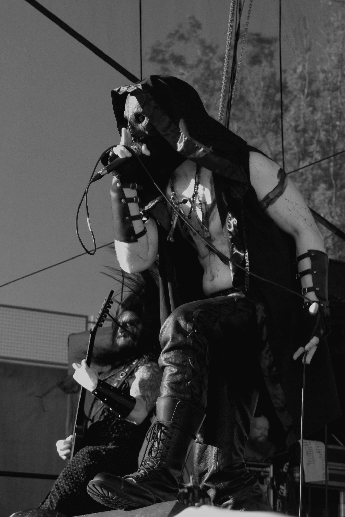Czarno-biała fotografia koncertowa: wokalista w masce i kapturze, z gitarzystą w tle, podczas występu na festiwalu MetalMine 2024. Dynamiczne ujęcie, ostre światło.