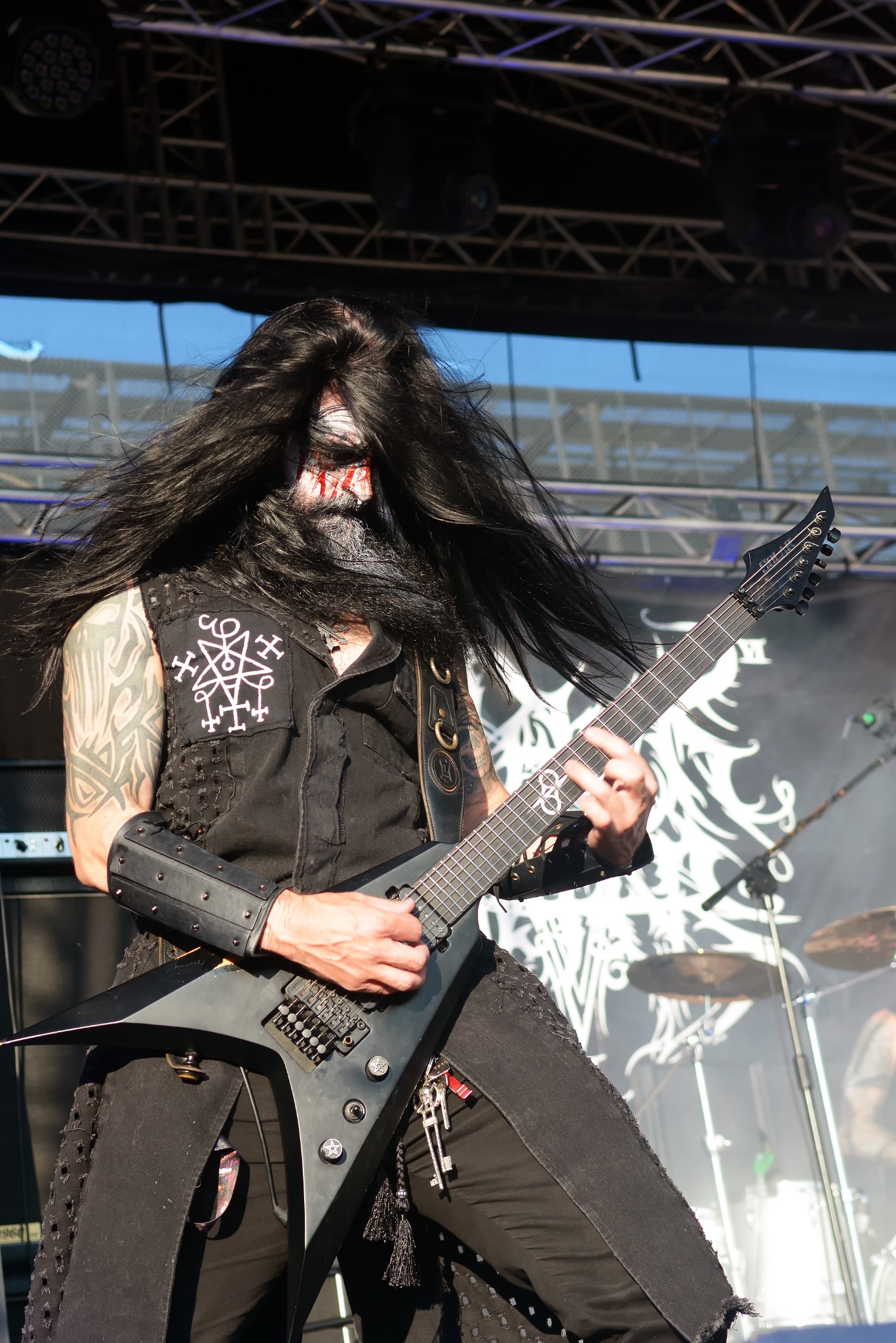 Koncertowy kadr: gitarzysta black metalowy z demonicznym makijażem gra na czarnej gitarze typu V na tle sceny z perkusją, oświetleniem i elementami dekoracji.