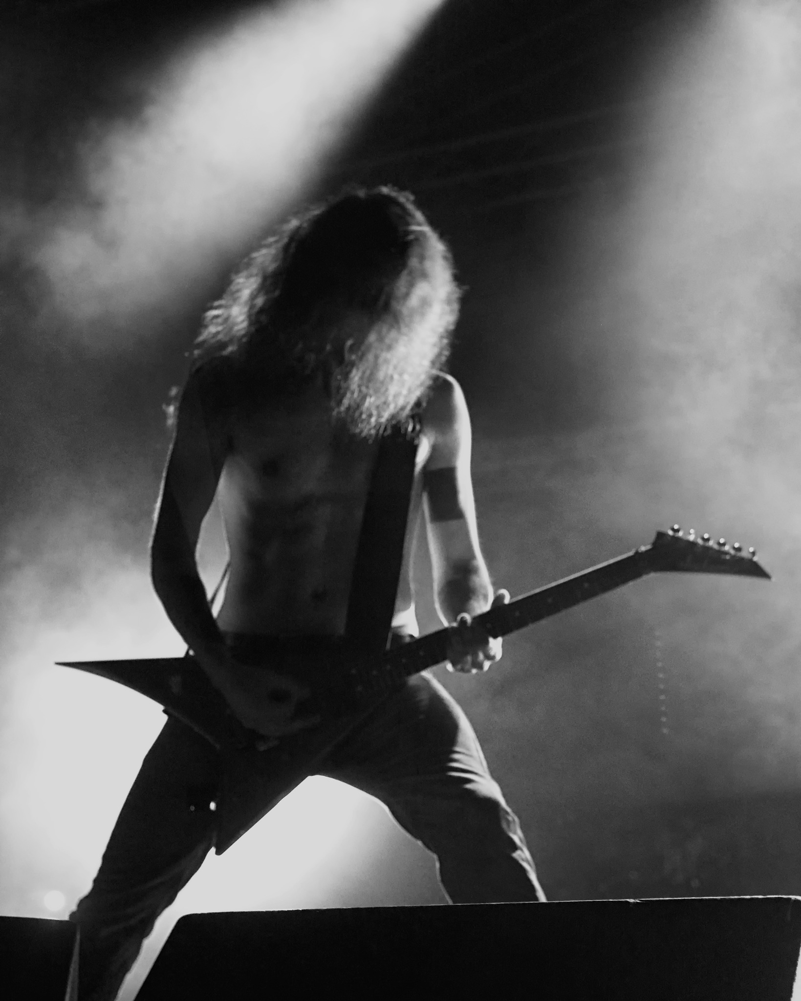 Czarno-białe zdjęcie z koncertu: gitarzysta gra na gitarze typu V na tle dymu i reflektorów. Widoczny tors i długie włosy muzyka.