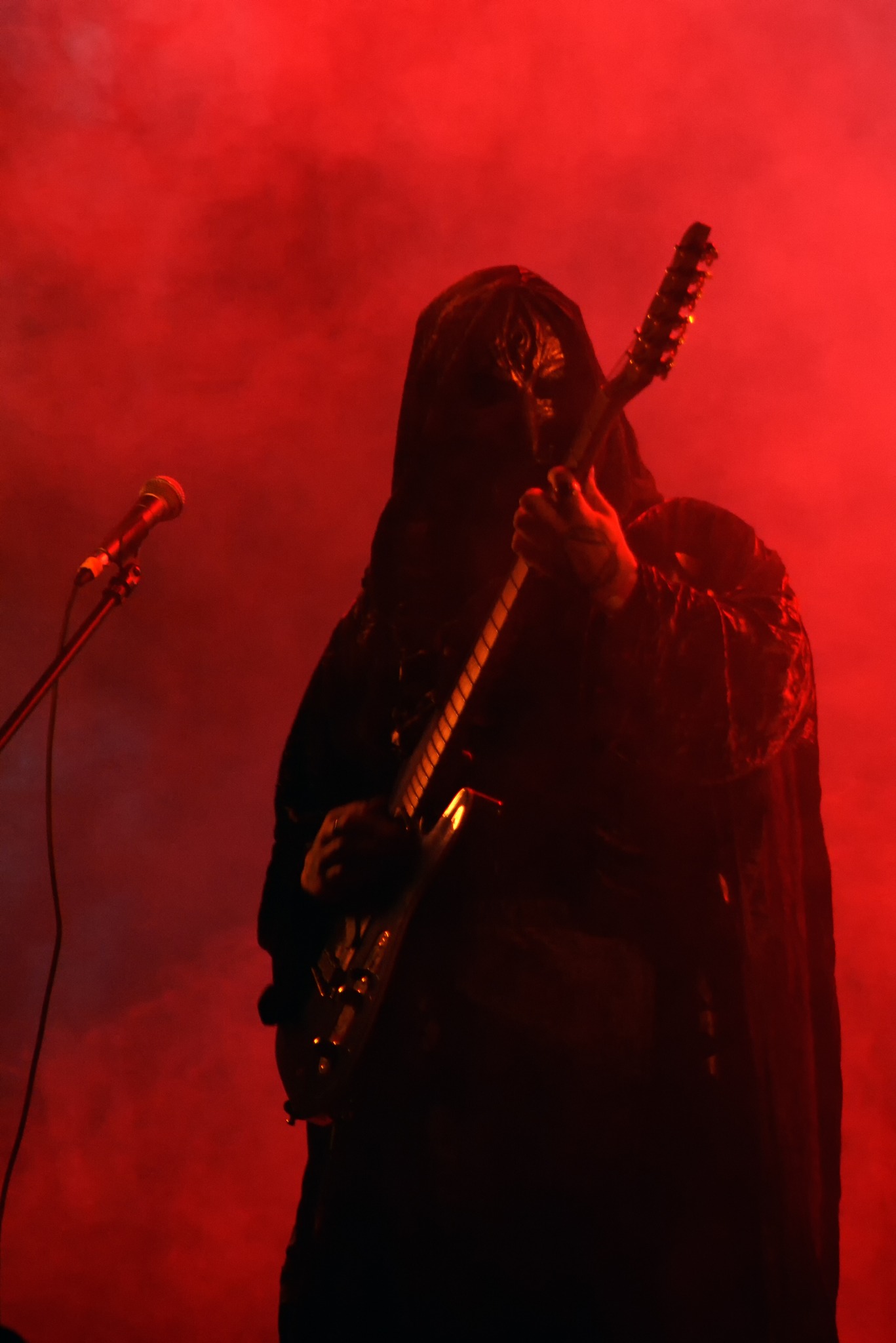 Koncertowy kadr: Zamaskowany gitarzysta w kapturze, otoczony czerwonym dymem i światłem, gra na gitarze elektrycznej obok mikrofonu. Mroczna scena muzyczna.