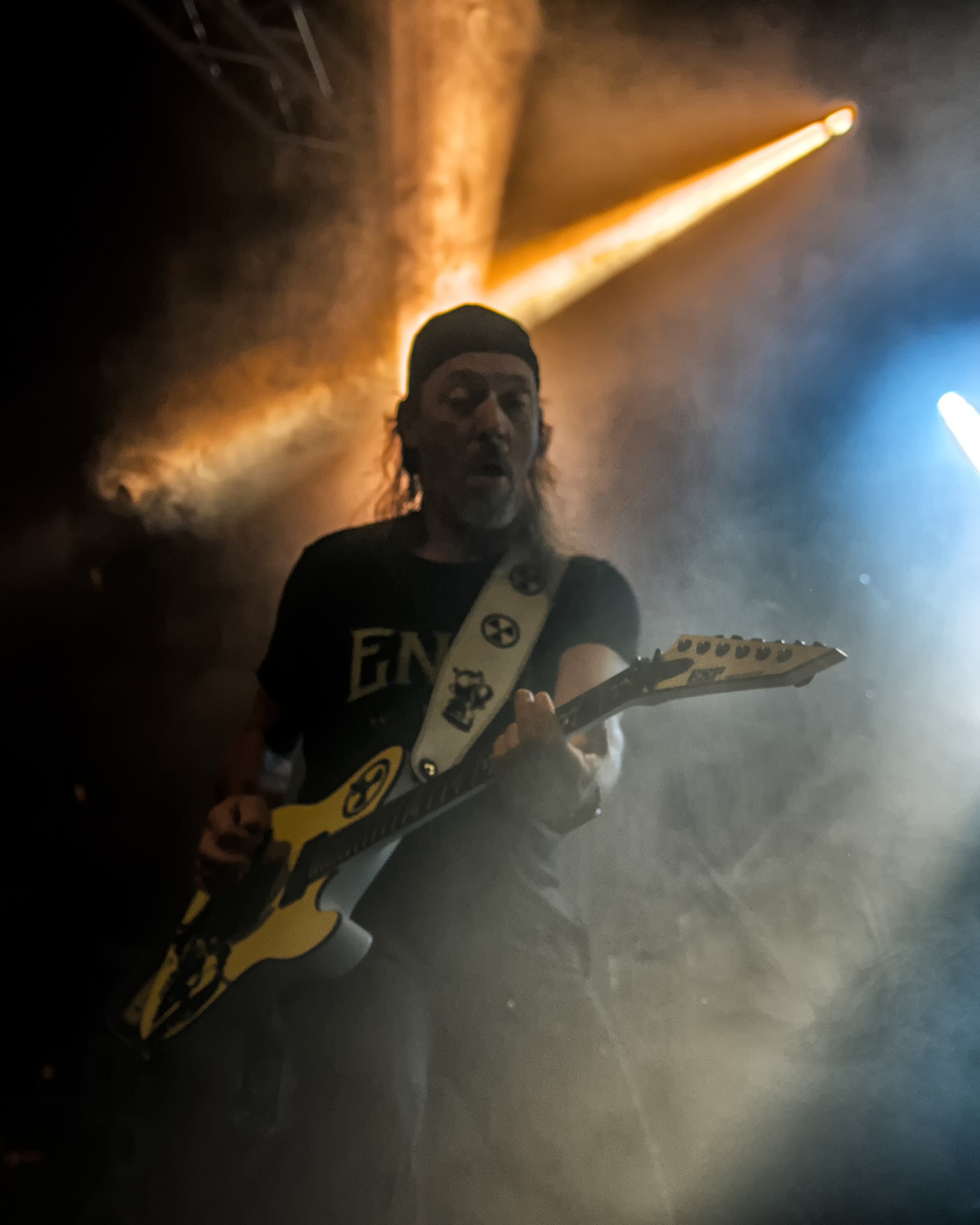 Gitarzysta z długimi włosami w czarnej koszulce gra na żółto-białej gitarze ESP na scenie w oświetleniu pomarańczowo-niebieskim i dymie.