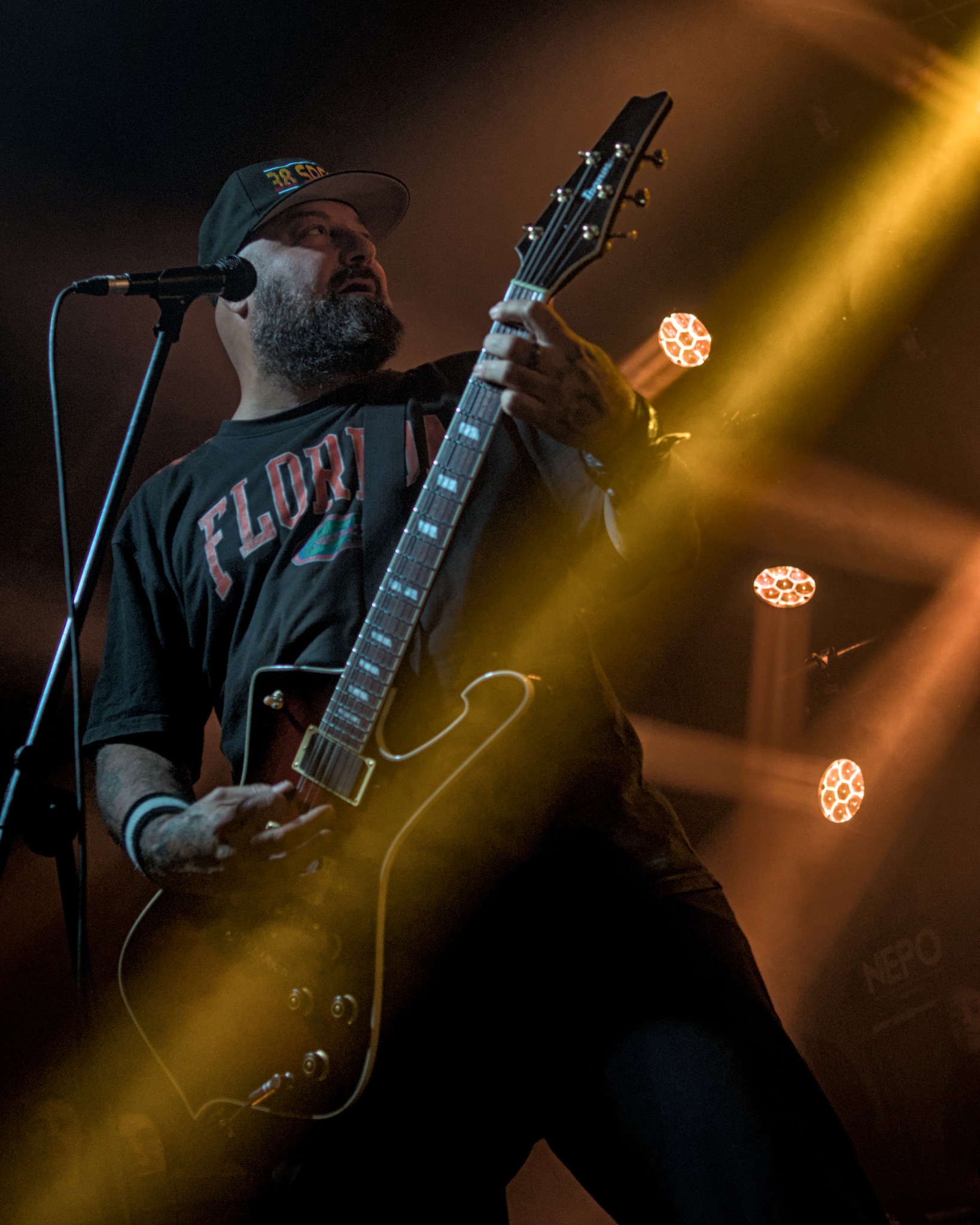 Gitarzysta z brodą w czarnej koszulce 'Florida' gra na gitarze elektrycznej podczas koncertu, oświetlony pomarańczowym światłem reflektorów.