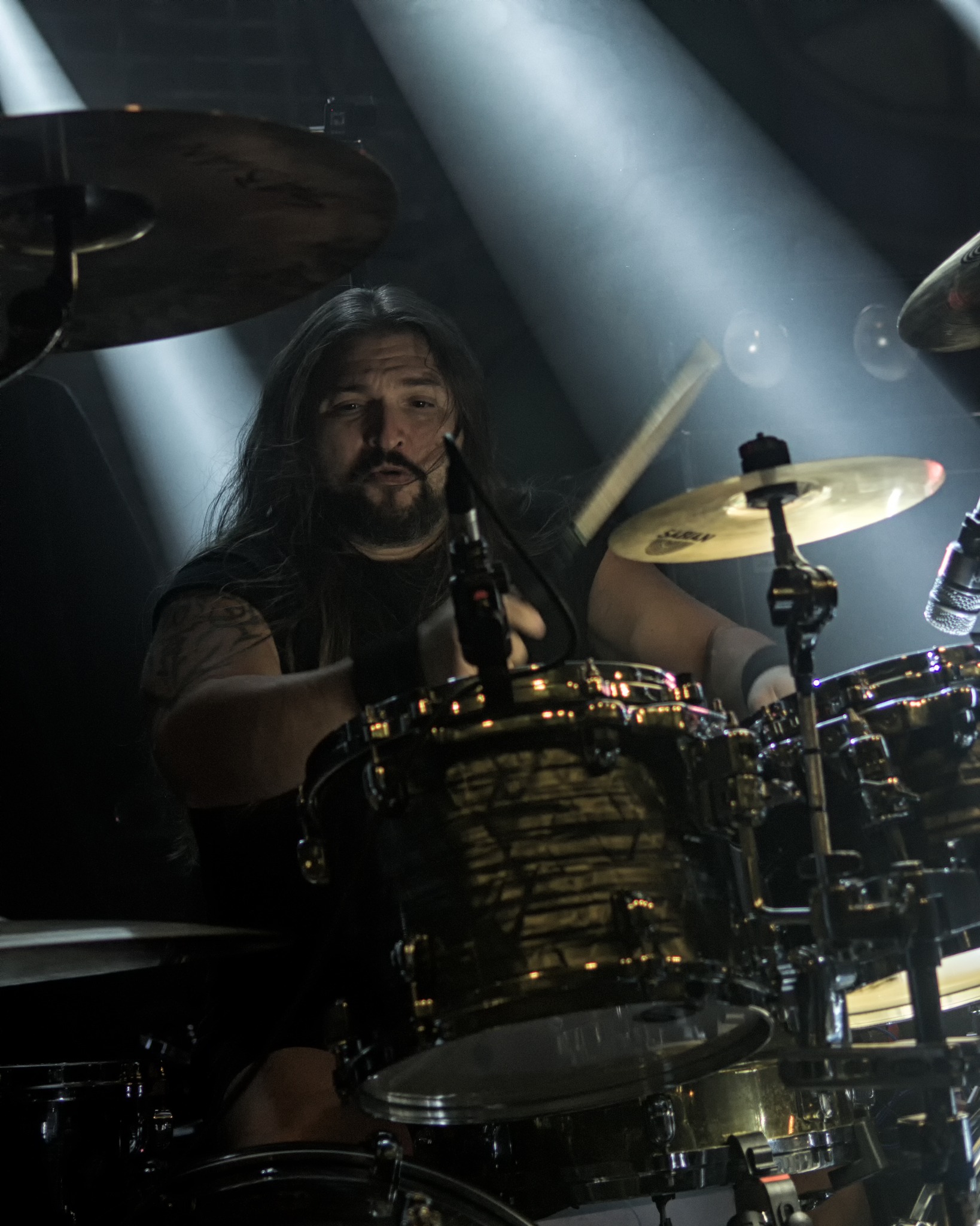 Perkusista z długimi włosami gra na perkusji pod światłami reflektorów. Widoczny tatuaż na ramieniu i mikrofon przed perkusją. Dynamiczne ujęcie z koncertu.