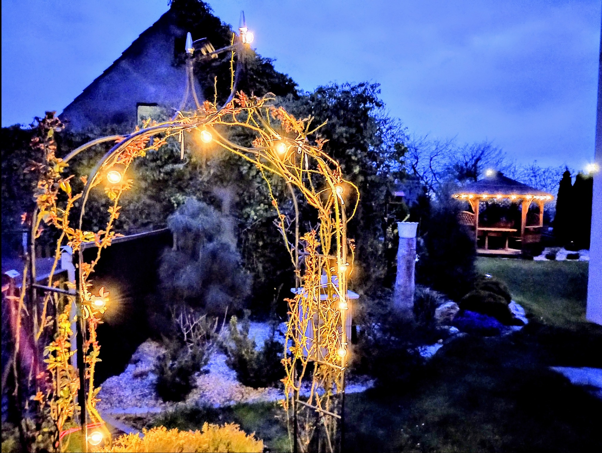 Oświetlony łuk z roślinami i lampkami w ogrodzie, w tle widoczna altana i dom o zmierzchu.