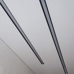 Serhii Holub S-Bud-Rem - Dwa równoległe, liniowe panele LED z czarnymi obramowaniami zamontowane na białym suficie podwieszanym w pomieszczeniu z widoczną fakturą ściany i fragmentem drewnianych drzwi.