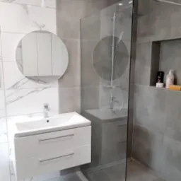 Nowoczesna łazienka z kabiną prysznicową typu walk-in, umywalką z szafką wiszącą i okrągłym lustrem, wykończona płytkami w odcieniach szarości i bieli z marmurkowym wzorem.