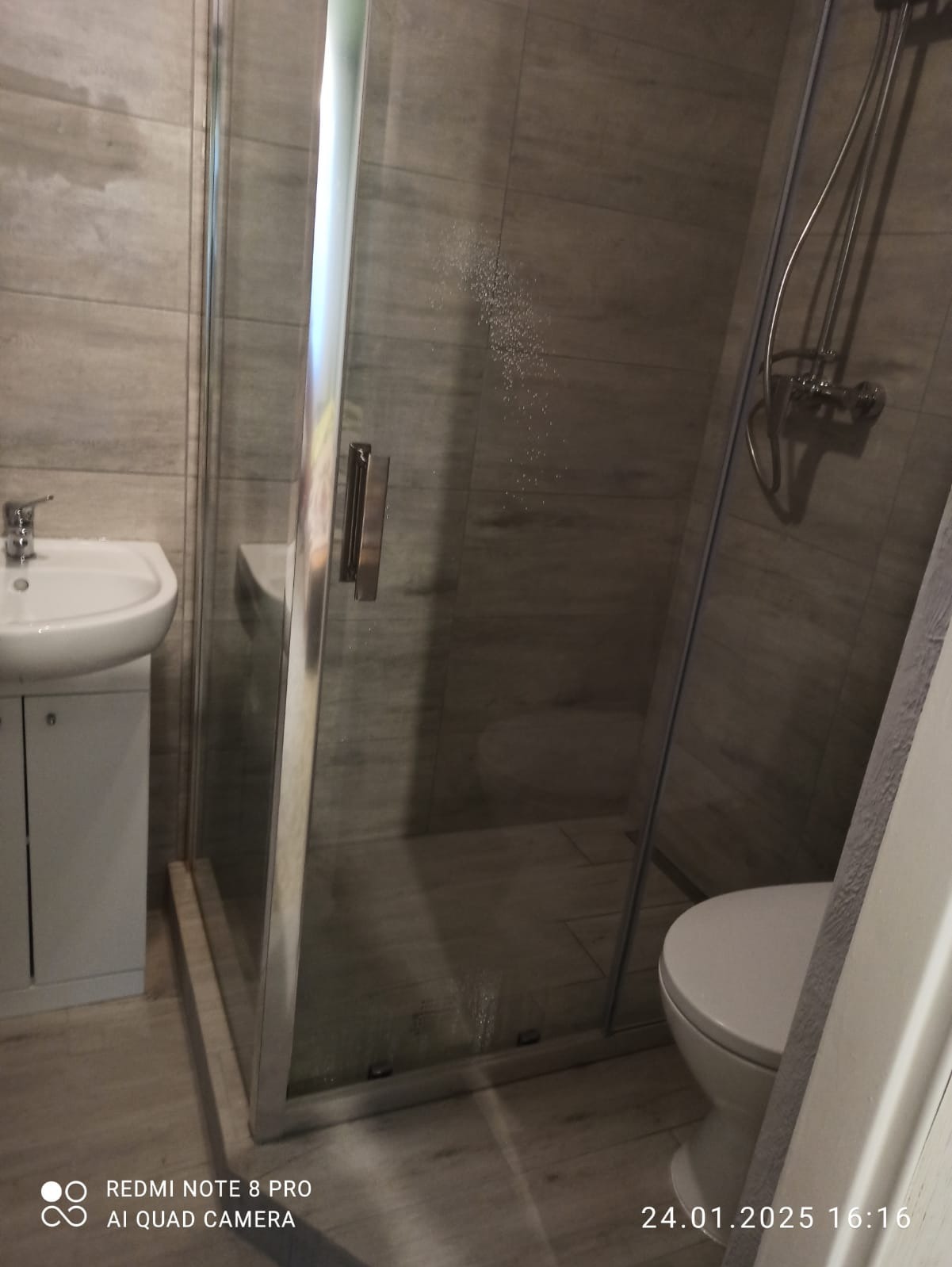 Nowoczesna łazienka z kabiną prysznicową typu walk-in, umywalką z szafką i toaletą, wykończona płytkami imitującymi drewno w odcieniach szarości.