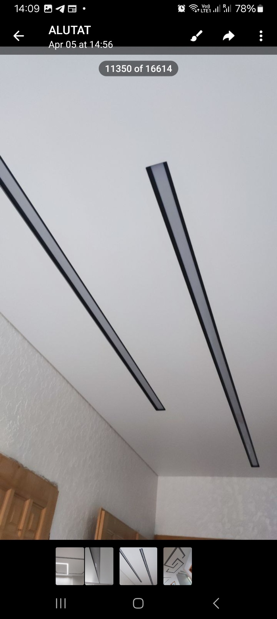 Dwa równoległe, liniowe panele LED z czarnymi obramowaniami zamontowane na białym suficie podwieszanym w pomieszczeniu z widoczną fakturą ściany i fragmentem drewnianych drzwi.