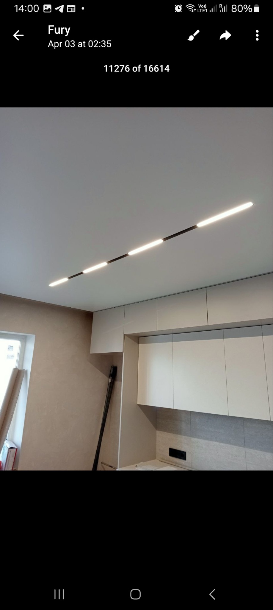 Nowoczesne oświetlenie sufitowe LED w kształcie linii, zamontowane w jasnym wnętrzu z zabudową kuchenną i ścianą w odcieniach beżu.