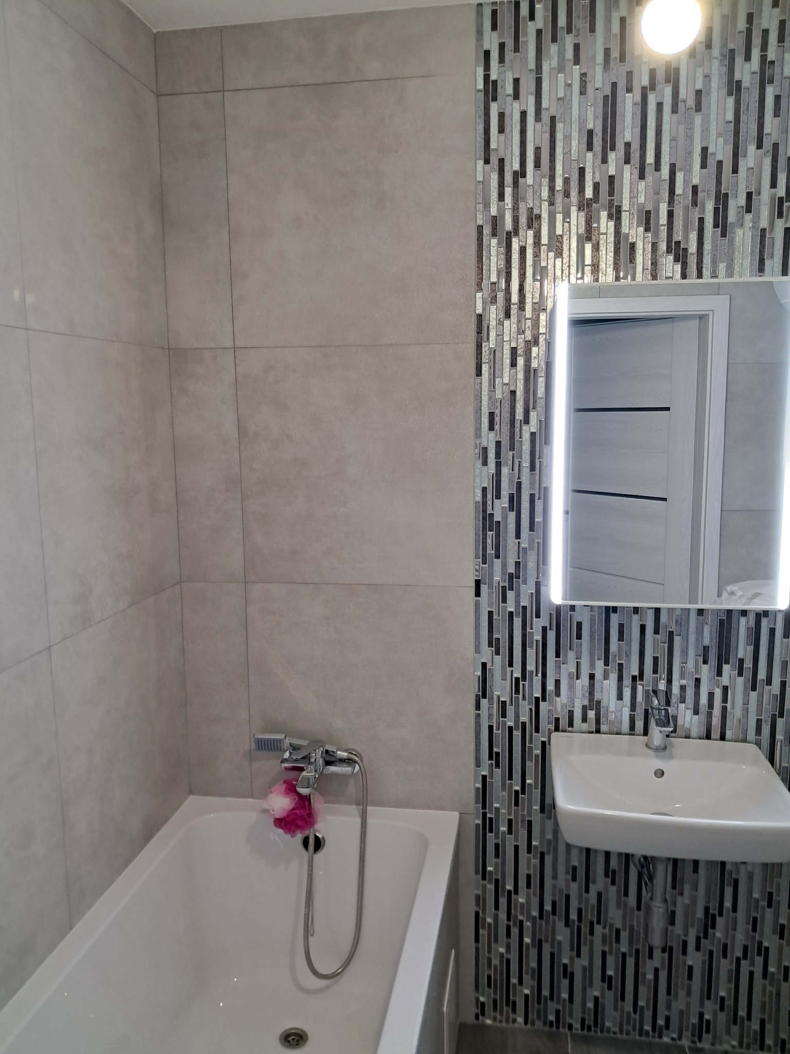 Nowoczesna łazienka z wanną, umywalką i podświetlanym lustrem, ściana wyłożona mozaiką w odcieniach szarości i srebra, na baterii wanny różowa myjka.