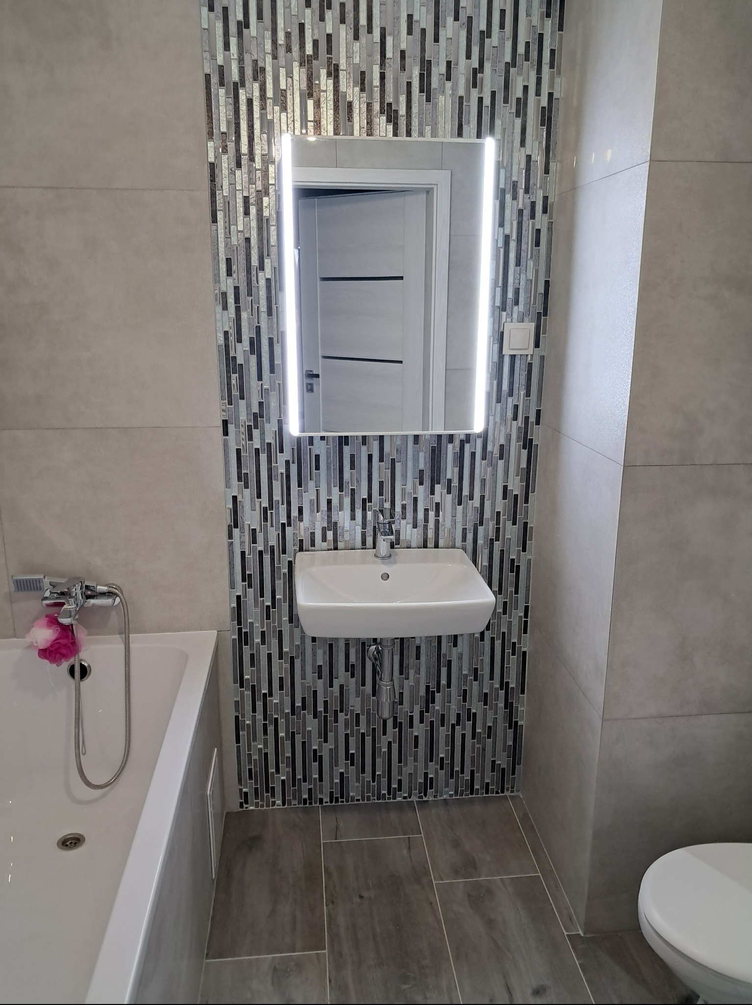 Nowoczesna łazienka z umywalką, podświetlanym lustrem otoczonym mozaiką i szarą glazurą.