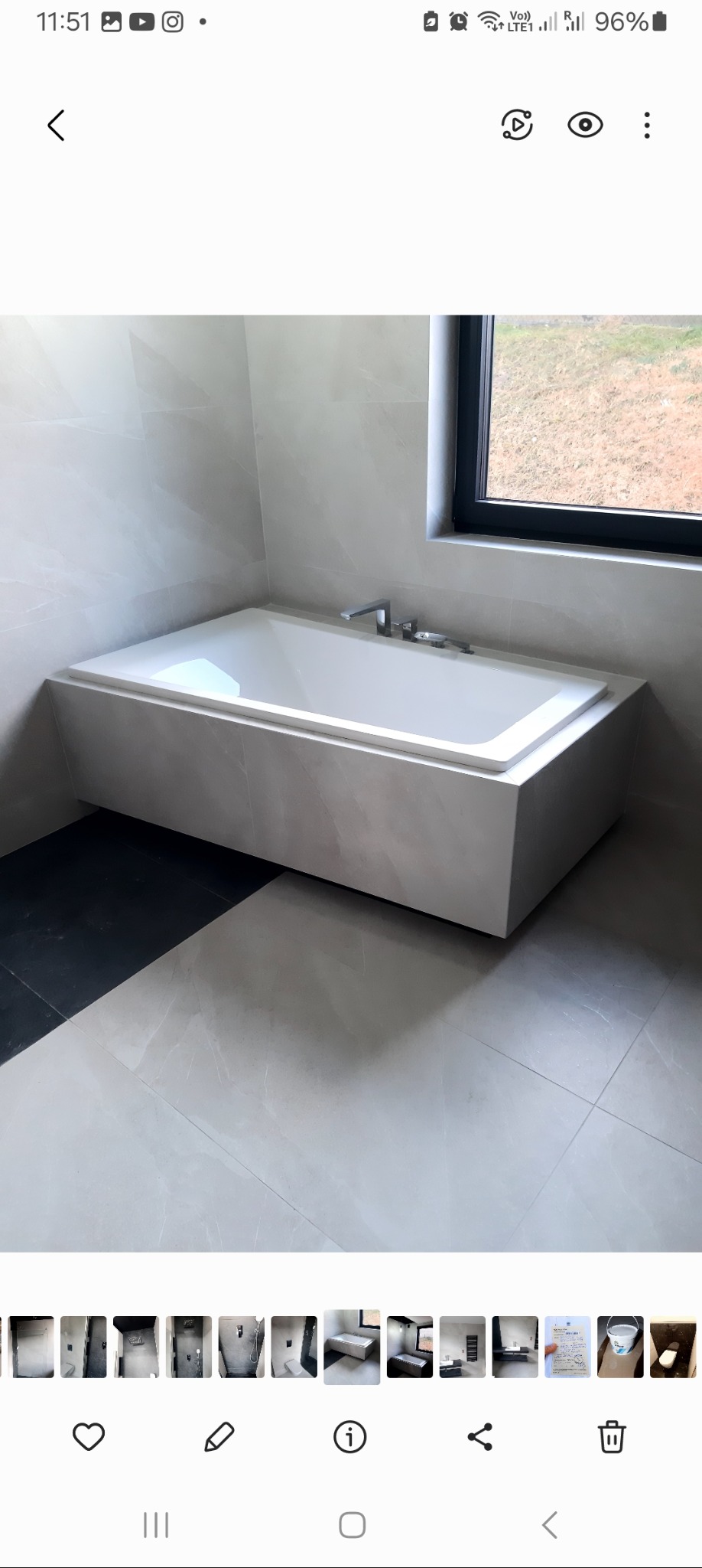 Nowoczesna wanna obudowana jasnoszarymi płytami imitującymi kamień, umieszczona w minimalistycznej łazience z oknem, wykończonej płytkami w odcieniach szarości i czerni.