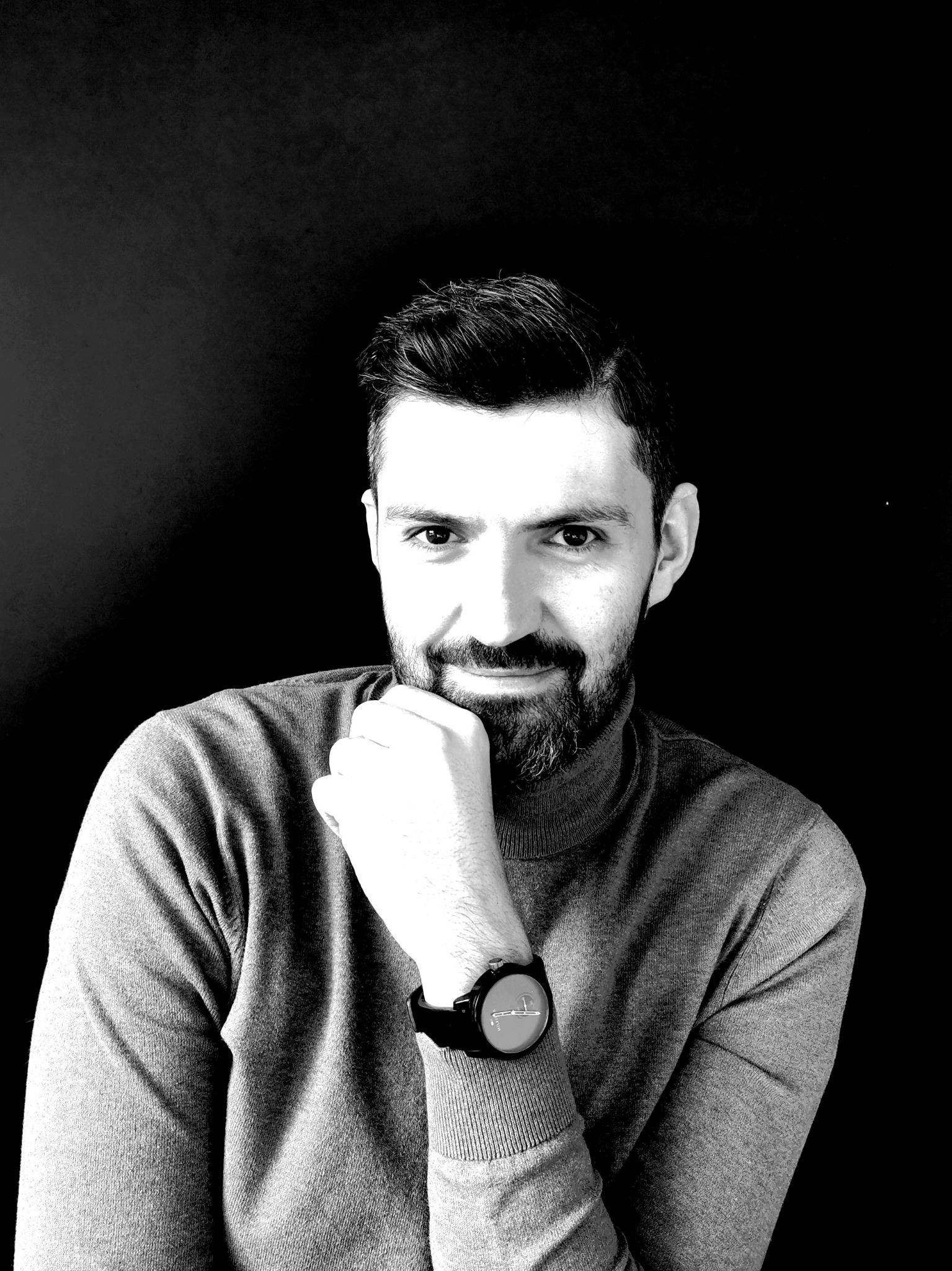 Portret mężczyzny z brodą w szarym golfie, opierającego podbródek na dłoni z zegarkiem na czarnym tle.