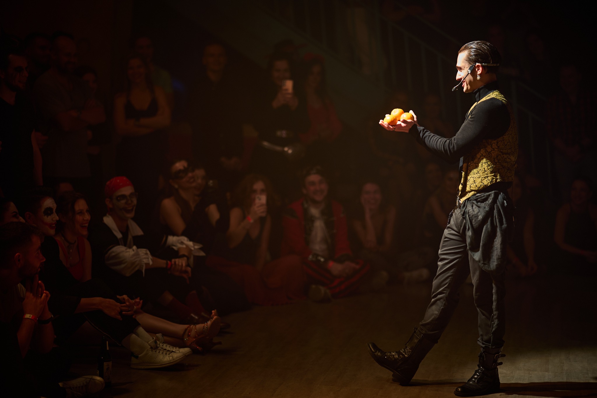 Mężczyzna w czarnym golfie i złotej kamizelce, z mikrofonem nagłownym, żongluje trzema pomarańczami na dłoni, na tle publiczności w kostiumach.