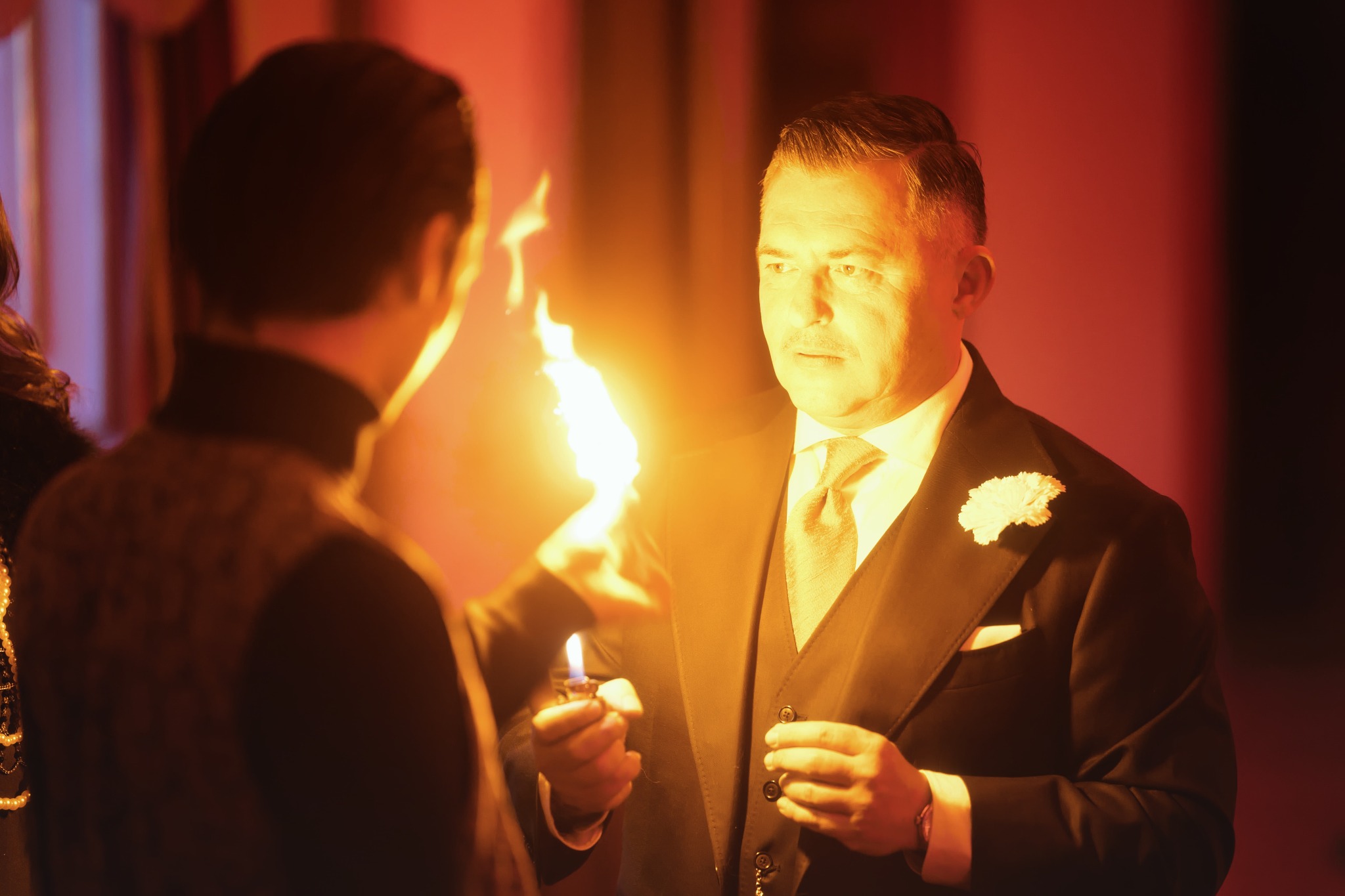 Iluzjonista w smokingu z butonierką, zapala ogień z zapalniczki przed widzem, tworząc magiczny efekt podczas występu.