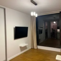 Nowoczesne wnętrze pokoju z parkietem, wiszącym telewizorem, małą półką, przesuwną szafą, zasłoniętym oknem balkonowym i lampą wiszącą z trzema białymi kloszami.