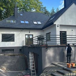 RIKO GROUP SPÓŁKA Z OGRANICZONĄ ODPOWIEDZIALNOŚCIĄ - Pracownik w kamizelce maluje elewację domu z czarnym dachem i balkonem, widoczne puszki z farbą, drabina i schody z ażurowych płyt.