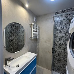 RIKO GROUP SPÓŁKA Z OGRANICZONĄ ODPOWIEDZIALNOŚCIĄ - Nowoczesna łazienka z umywalką na niebieskiej szafce, okrągłym lustrem i kabiną prysznicową z motywem liści. Grzejnik drabinkowy na ścianie. Jasne płytki.