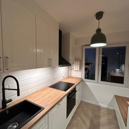 RIKO GROUP SPÓŁKA Z OGRANICZONĄ ODPOWIEDZIALNOŚCIĄ - Nowoczesna kuchnia z białymi szafkami, drewnianym blatem i czarną armaturą. Uwagę przykuwa zielona lampa wisząca nad oknem z widokiem na nocne miasto.