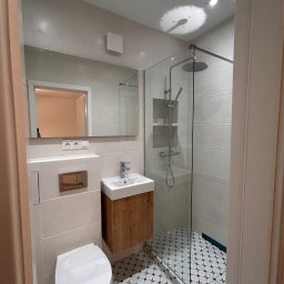 RIKO GROUP SPÓŁKA Z OGRANICZONĄ ODPOWIEDZIALNOŚCIĄ - Nowoczesna łazienka po remoncie: białe płytki, kabina prysznicowa ze szklaną ścianą, umywalka z drewnianą szafką i toaleta. Czarno-biała mozaika na podłodze.