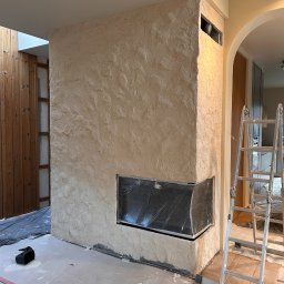 RIKO GROUP SPÓŁKA Z OGRANICZONĄ ODPOWIEDZIALNOŚCIĄ - Wykończony kominek z dekoracyjną, beżową strukturą tynku, zabezpieczony wkład kominkowy folią, drabina oparta o ścianę. Widoczne ślady prac wykończeniowych.