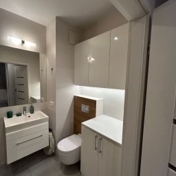 RIKO GROUP SPÓŁKA Z OGRANICZONĄ ODPOWIEDZIALNOŚCIĄ - Nowoczesna łazienka w bieli i szarości: umywalka z szafką, toaleta, szafki wiszące z podświetleniem LED, szare płytki podłogowe. Minimalistyczny design.