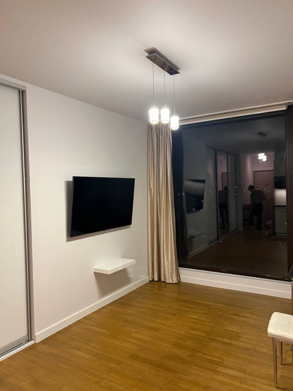 Nowoczesne wnętrze pokoju z parkietem, wiszącym telewizorem, małą półką, przesuwną szafą, zasłoniętym oknem balkonowym i lampą wiszącą z trzema białymi kloszami.