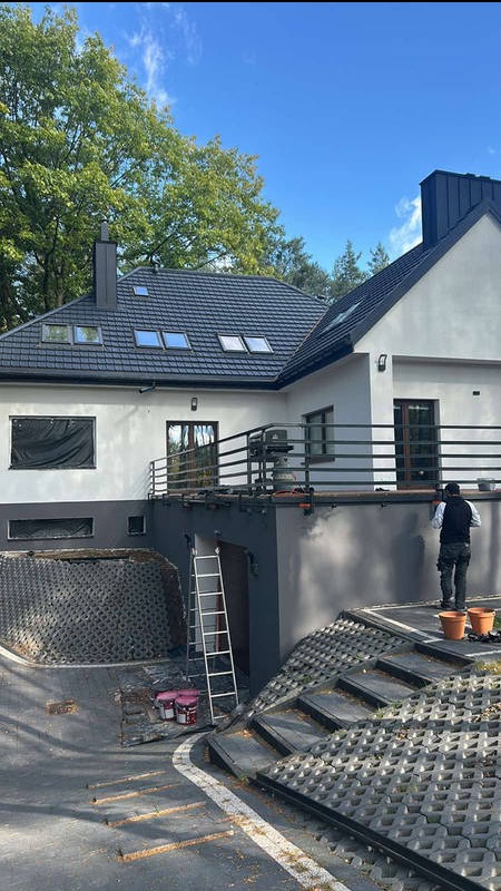 Pracownik w kamizelce maluje elewację domu z czarnym dachem i balkonem, widoczne puszki z farbą, drabina i schody z ażurowych płyt.