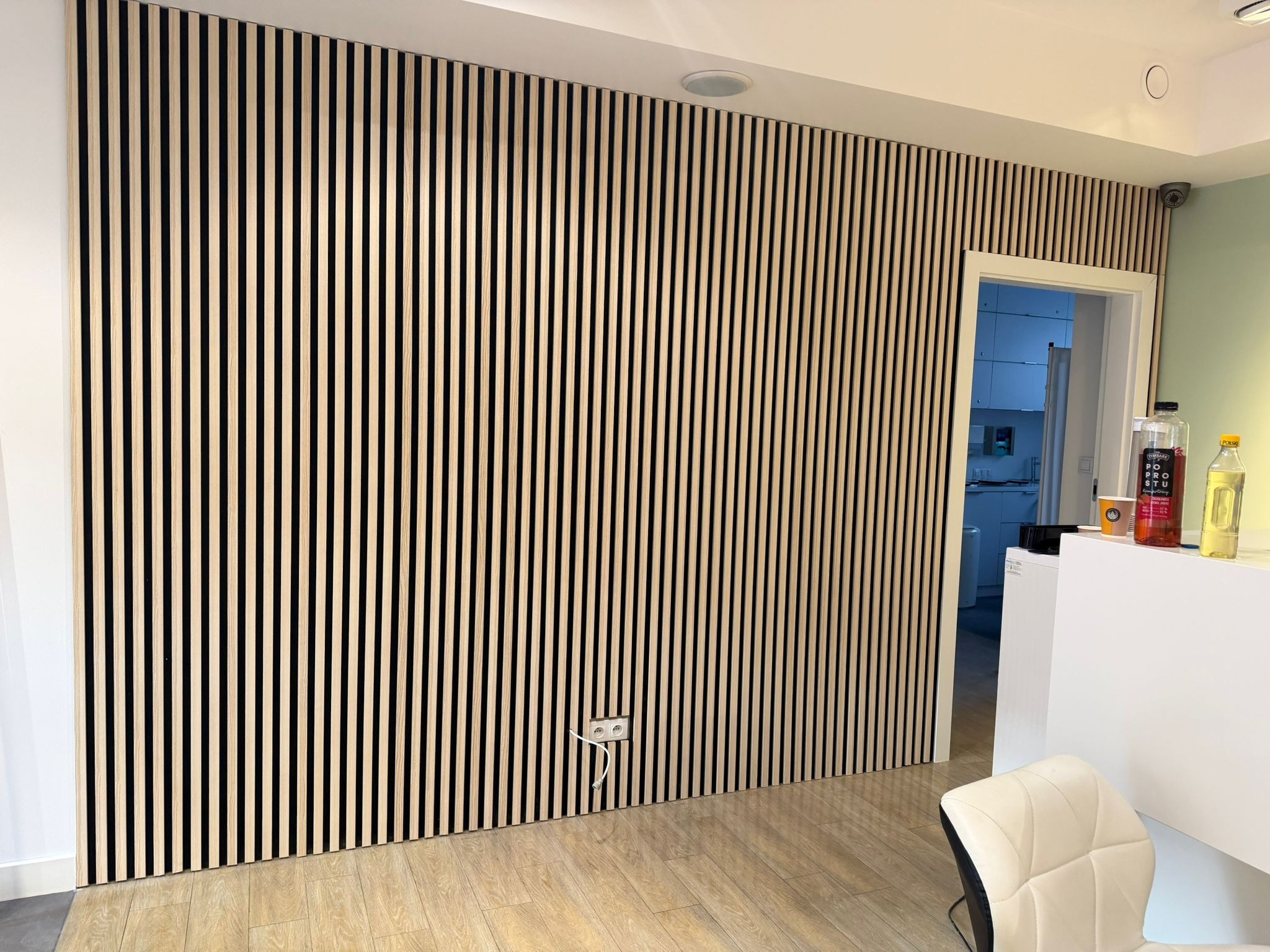 Ściana wykończona panelami lamelowymi w odcieniach beżu i czerni, z widocznym gniazdkiem elektrycznym i fragmentem wnętrza z białymi meblami w tle.