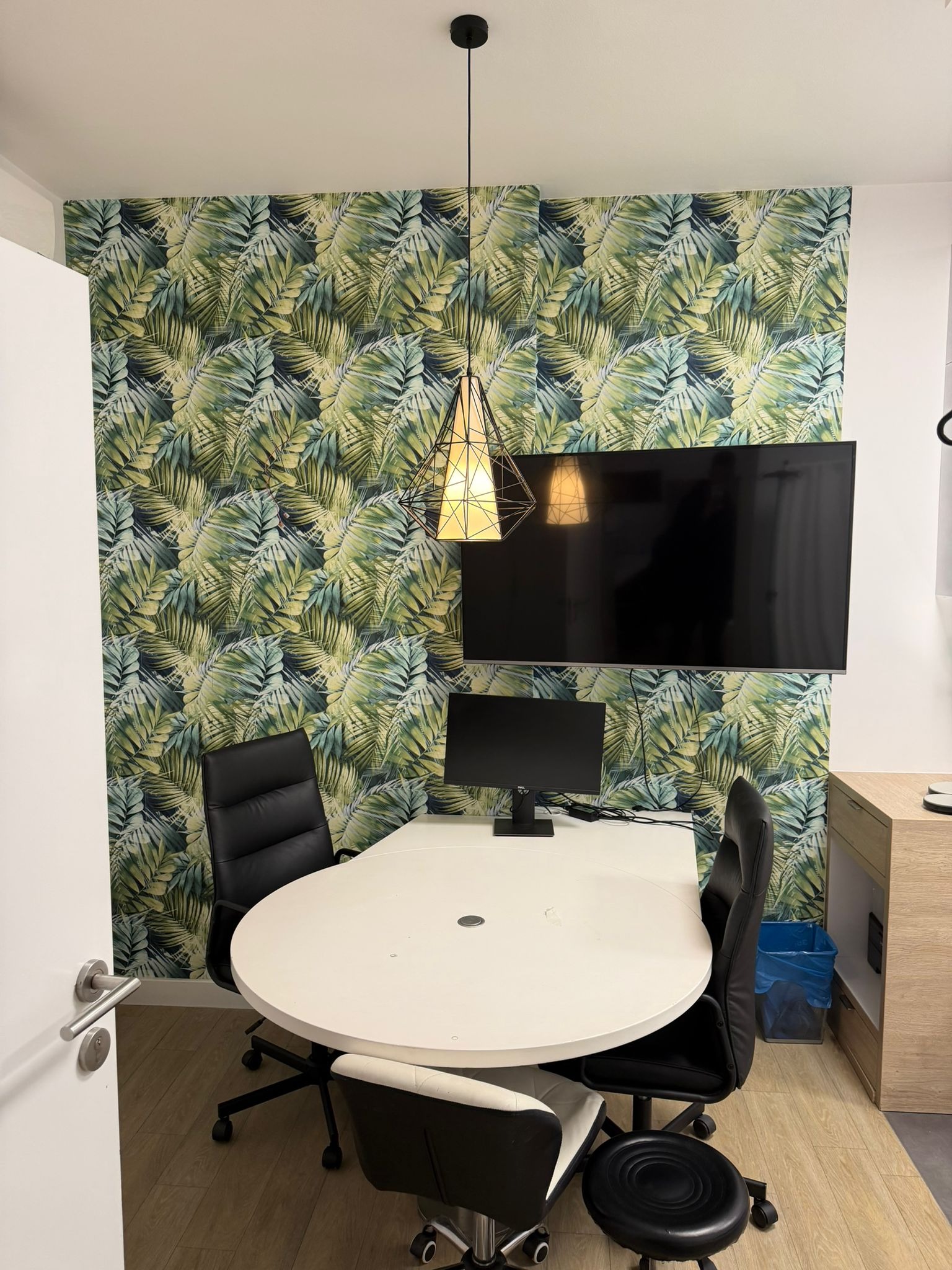 Nowoczesne biuro z okrągłym stołem, krzesłami i ścianą w tropikalne liście. Telewizor i lampa wisząca dodają charakteru. Widok na odnowione wnętrze biura.