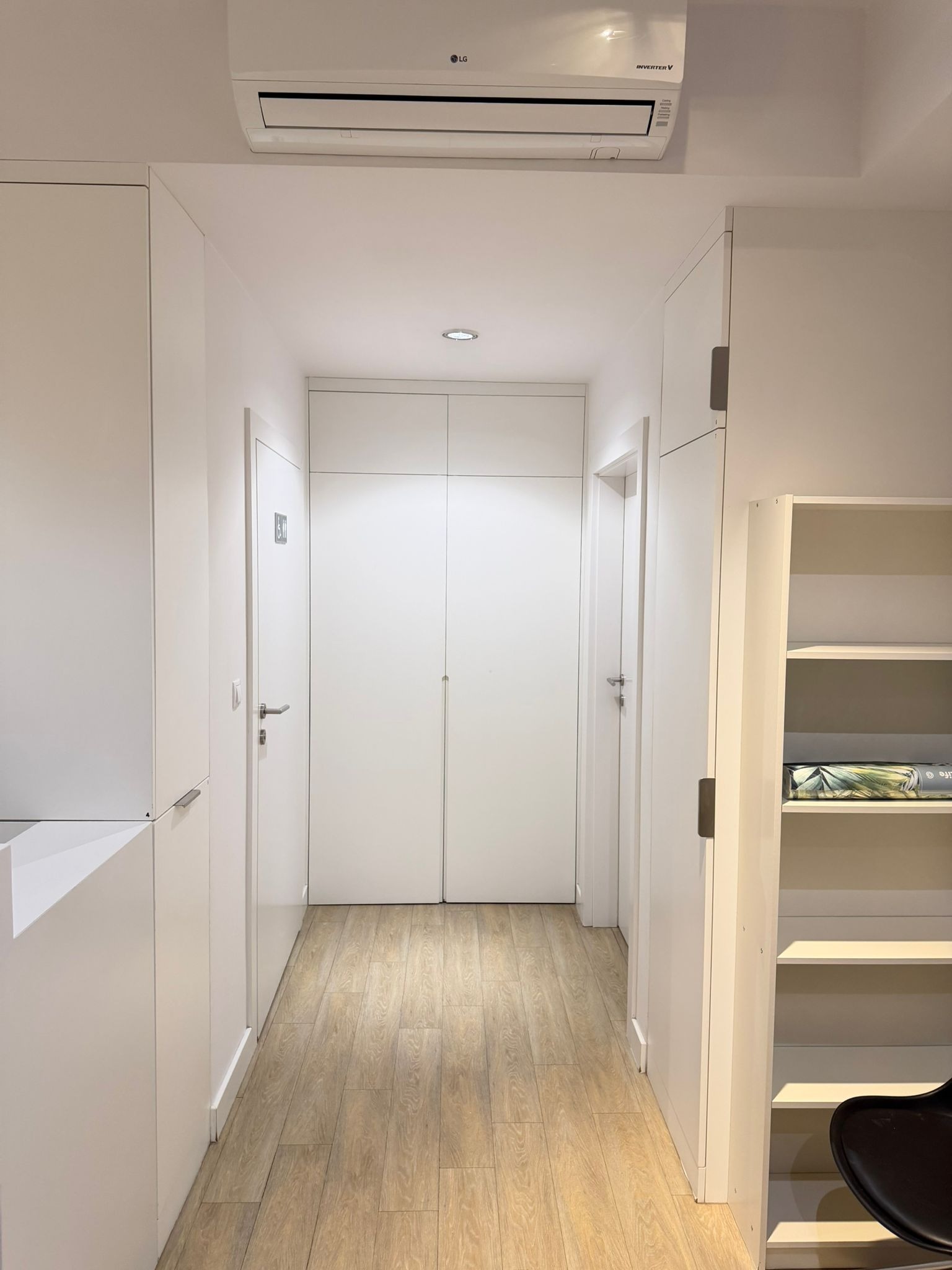 Białe, minimalistyczne wnętrze: korytarz z drewnianą podłogą, zabudowane szafy, drzwi i klimatyzacja LG. Nowoczesny design, czyste linie, jasne oświetlenie.