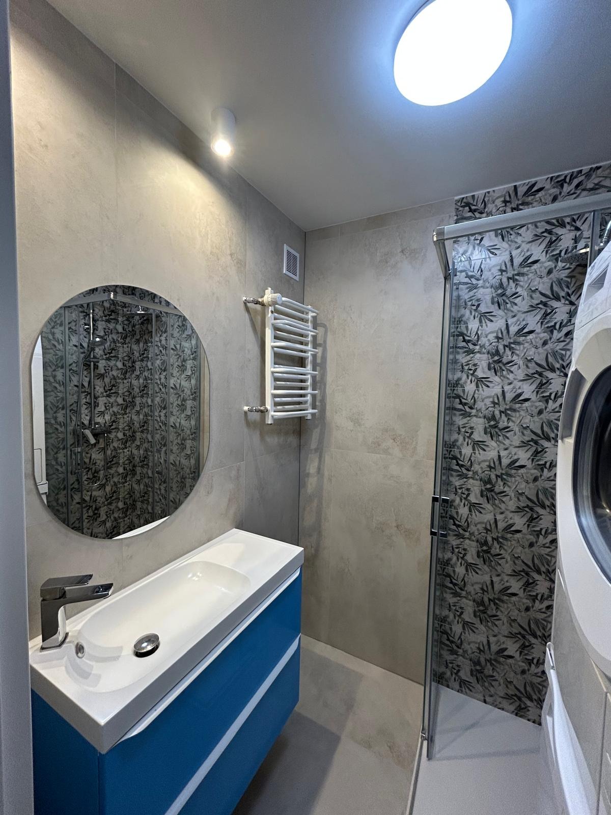 Nowoczesna łazienka z umywalką na niebieskiej szafce, okrągłym lustrem i kabiną prysznicową z motywem liści. Grzejnik drabinkowy na ścianie. Jasne płytki.