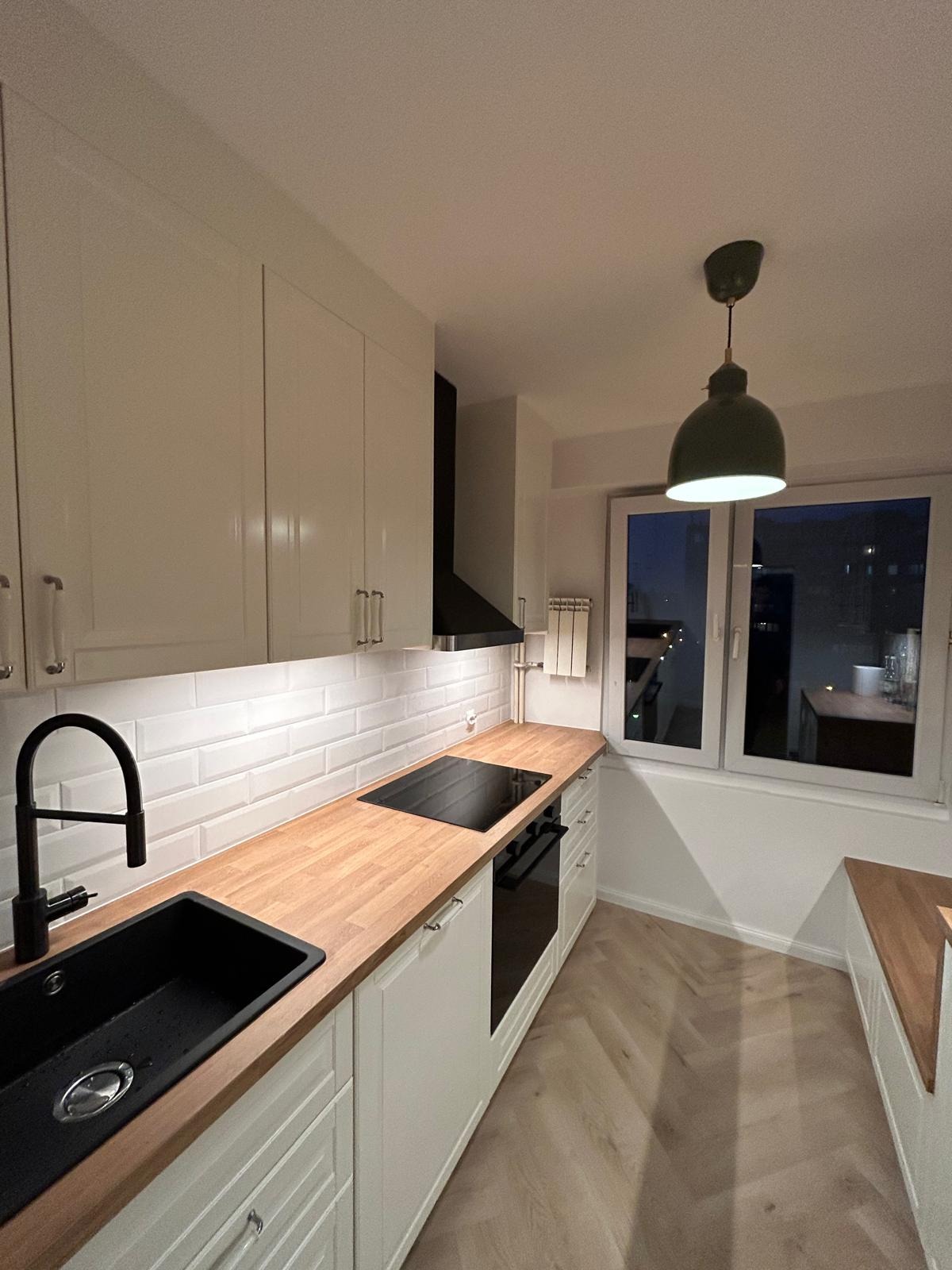 Nowoczesna kuchnia z białymi szafkami, drewnianym blatem i czarną armaturą. Uwagę przykuwa zielona lampa wisząca nad oknem z widokiem na nocne miasto.