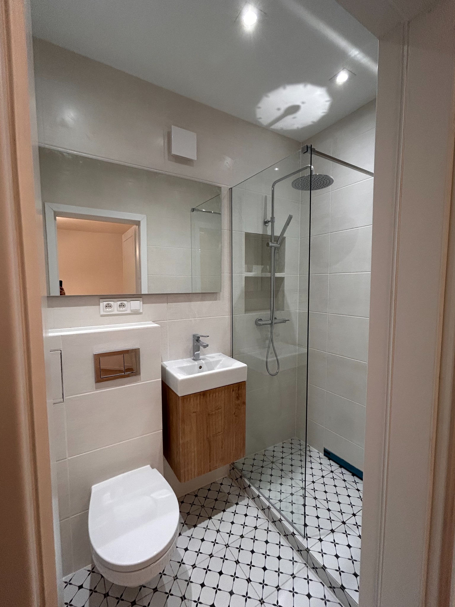 Nowoczesna łazienka po remoncie: białe płytki, kabina prysznicowa ze szklaną ścianą, umywalka z drewnianą szafką i toaleta. Czarno-biała mozaika na podłodze.