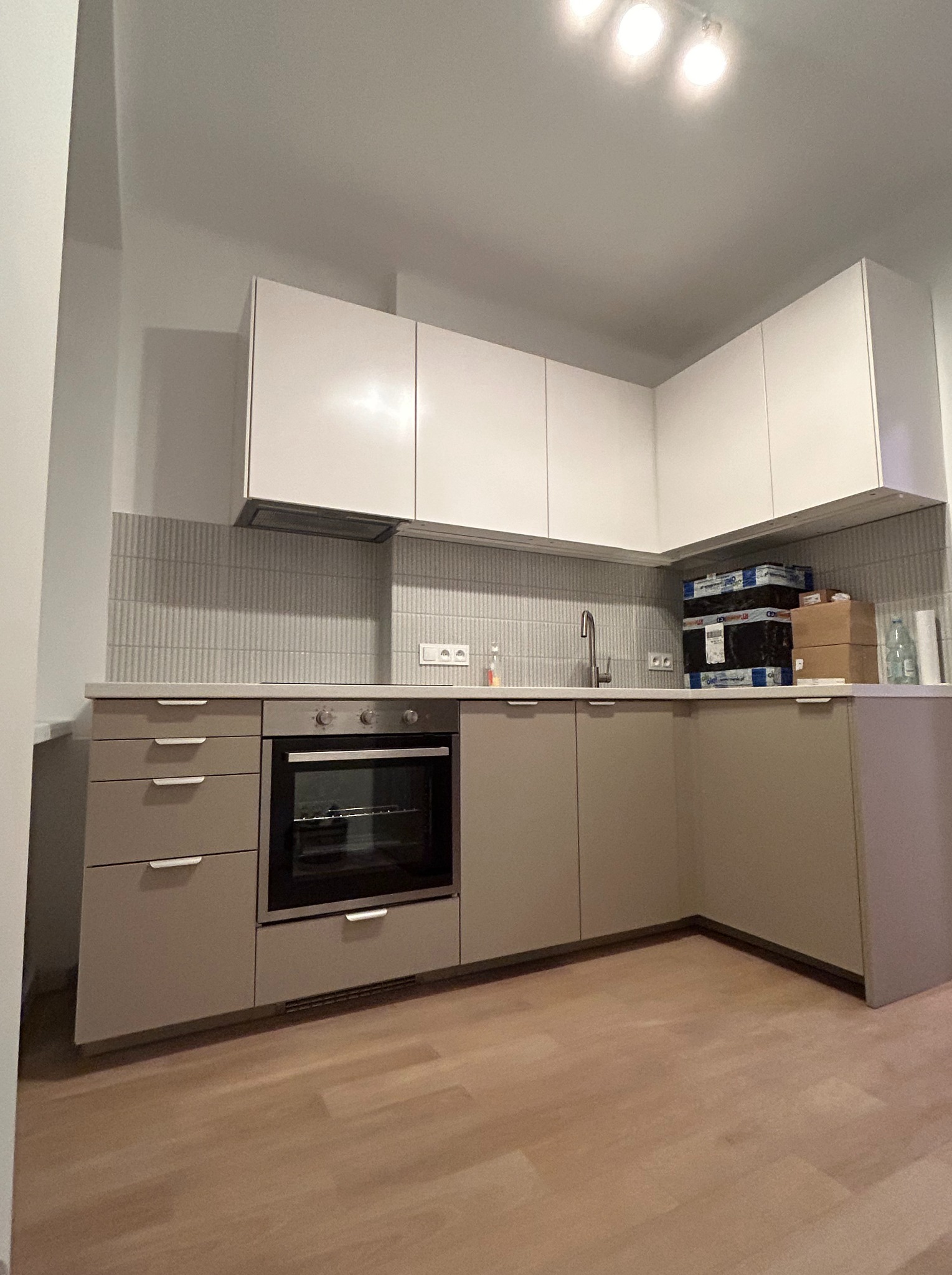 Nowoczesna kuchnia w odcieniach beżu i bieli. Widoczne szafki, piekarnik, płytki na ścianie i drewniana podłoga. Na blacie stoją kartony i butelka.