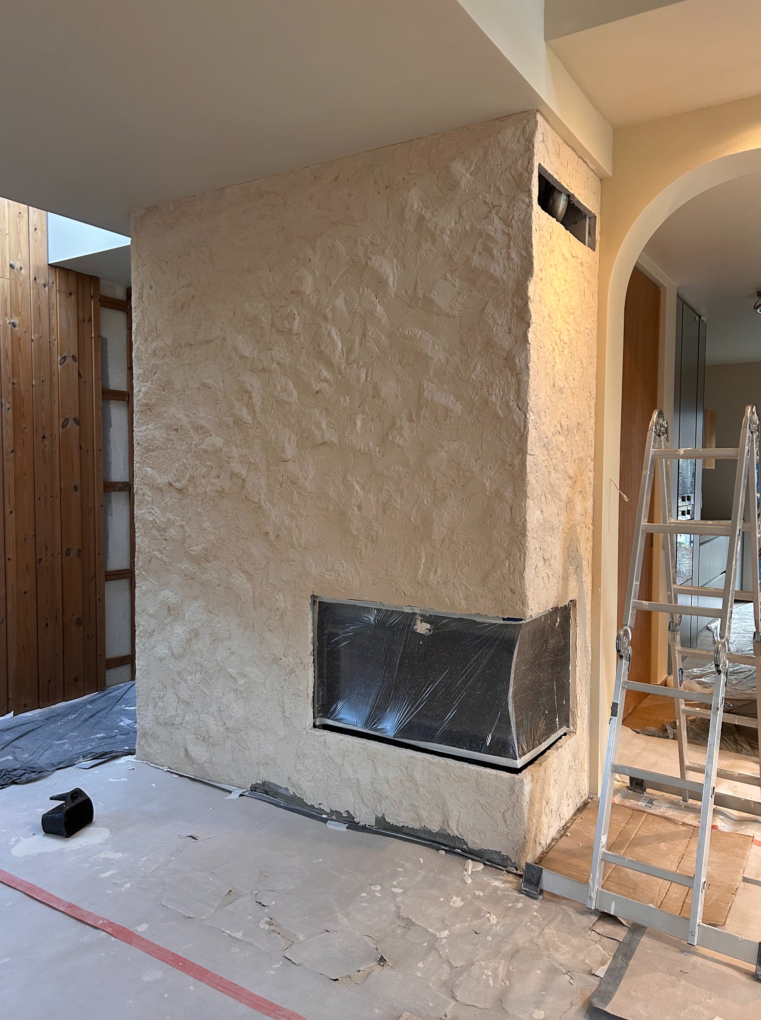 Wykończony kominek z dekoracyjną, beżową strukturą tynku, zabezpieczony wkład kominkowy folią, drabina oparta o ścianę. Widoczne ślady prac wykończeniowych.