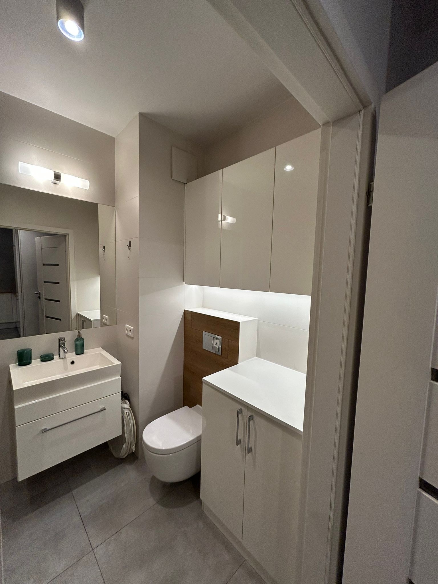 Nowoczesna łazienka w bieli i szarości: umywalka z szafką, toaleta, szafki wiszące z podświetleniem LED, szare płytki podłogowe. Minimalistyczny design.