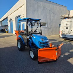 P&D OGRODY PIOTR BUDZYŃSKI - Niebieski traktor z pomarańczowym pługiem śnieżnym na tle białego budynku. Sprzęt gotowy do pracy, oświetlony jasnym słońcem. Widoczny fragment zaparkowanego busa.