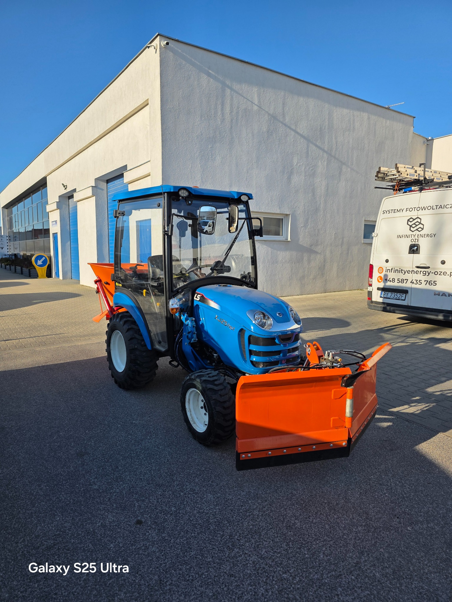 Niebieski traktor z pomarańczowym pługiem śnieżnym na tle białego budynku. Sprzęt gotowy do pracy, oświetlony jasnym słońcem. Widoczny fragment zaparkowanego busa.