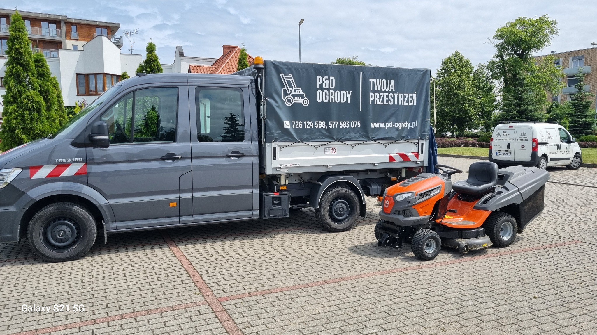 Szary samochód dostawczy z logo firmy ogrodniczej i pomarańczowa kosiarka samojezdna na kostce brukowej. W tle budynki i zieleń. Drugi samochód firmy w oddali.