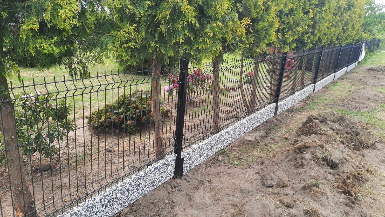 Czarny panelowy płot z podmurówką z szarego kruszywa, oddzielający ogród z drzewami i kwiatami od terenu z ziemią i trawą.