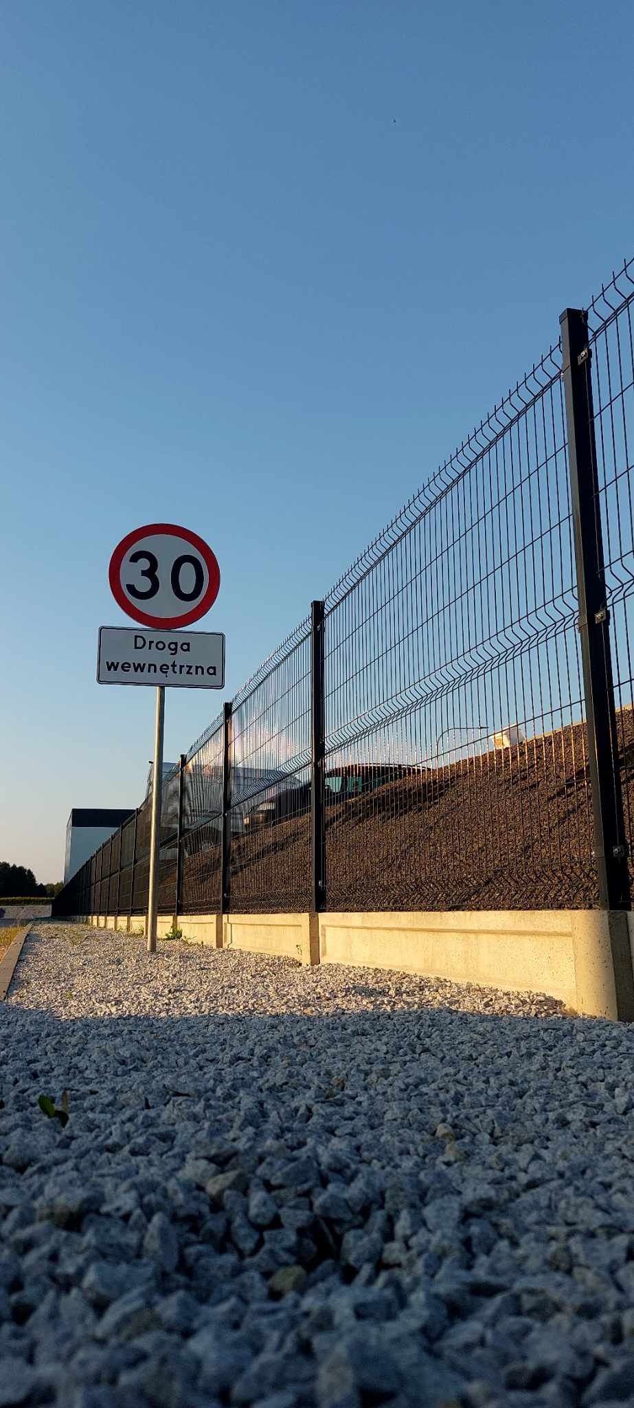 Nowoczesne ogrodzenie panelowe z siatki, czarne słupki, podmurówka betonowa, znak ograniczenia prędkości do 30 km/h, droga wewnętrzna, żwirowe podłoże.