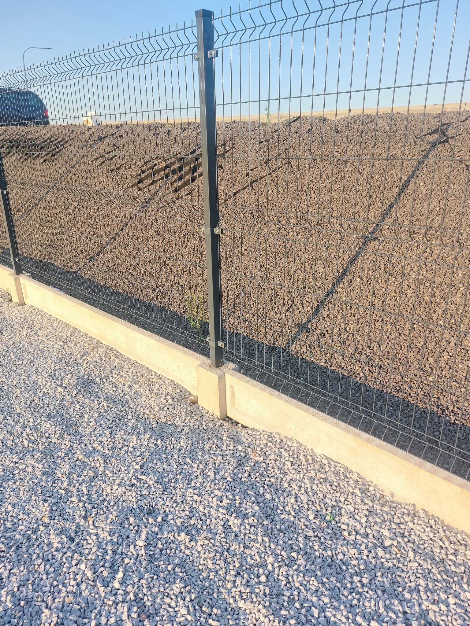 Szare ogrodzenie panelowe na betonowej podmurówce, z widocznym podłożem z białego żwiru i ziemnym nasypem w tle.