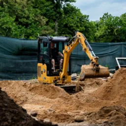 Operator w żółtej minikoparce CAT 301.8, nabierający piasek z wykopu na tle zieleni i ogrodzenia, z logo firmy budowlanej w prawym dolnym rogu.