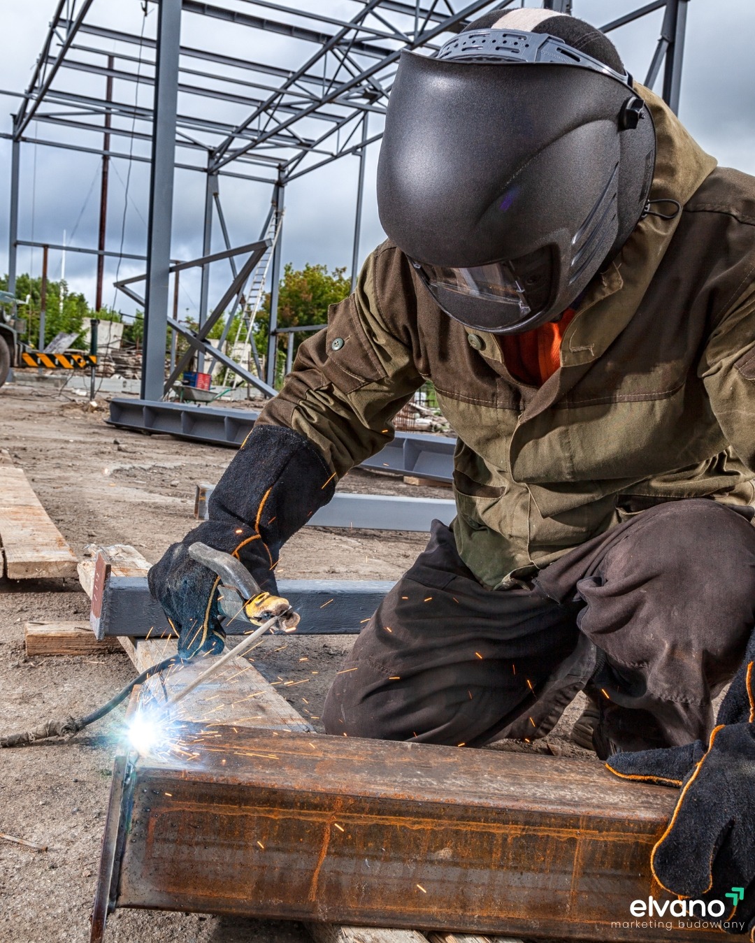Spawacz w ochronnym hełmie i rękawicach spawa element konstrukcyjny na placu budowy, widoczne iskry i metalowa konstrukcja w tle.