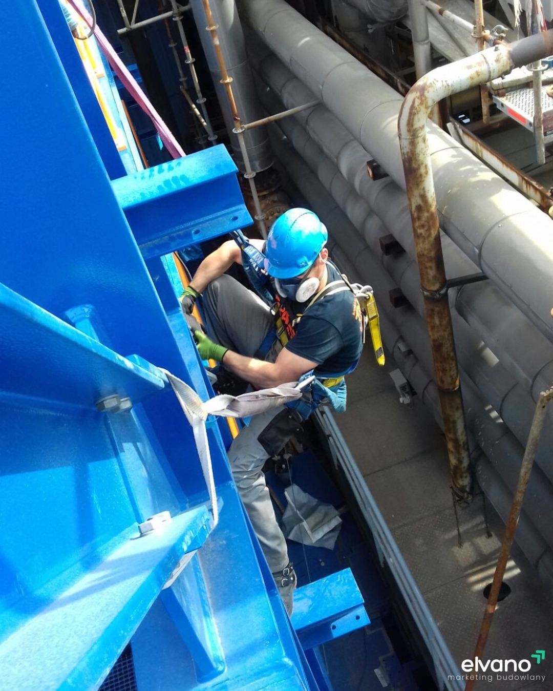Pracownik w kasku i masce ochronnej, zabezpieczony linami, maluje na niebiesko metalową konstrukcję na wysokości, widoczne rury i rusztowania w tle.