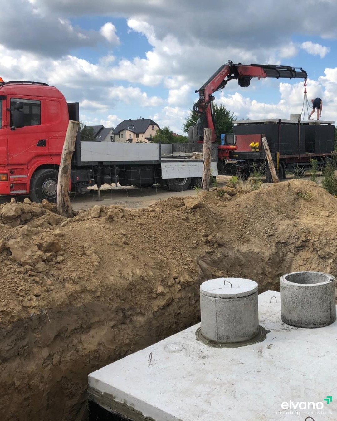 Montaż betonowych elementów kanalizacyjnych za pomocą dźwigu samochodowego na placu budowy, widoczny wykop i częściowo zakopana konstrukcja.