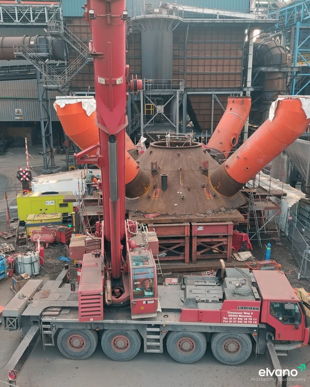 Czerwony dźwig Liebherr na terenie przemysłowym podczas montażu lub demontażu elementu konstrukcyjnego z trzema pomarańczowymi rurami, widok z góry.