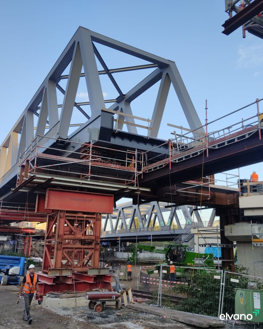 Konstrukcja stalowa mostu w trakcie budowy, widoczne rusztowania, pracownicy w kamizelkach odblaskowych i sprzęt budowlany, w tle drugi most.