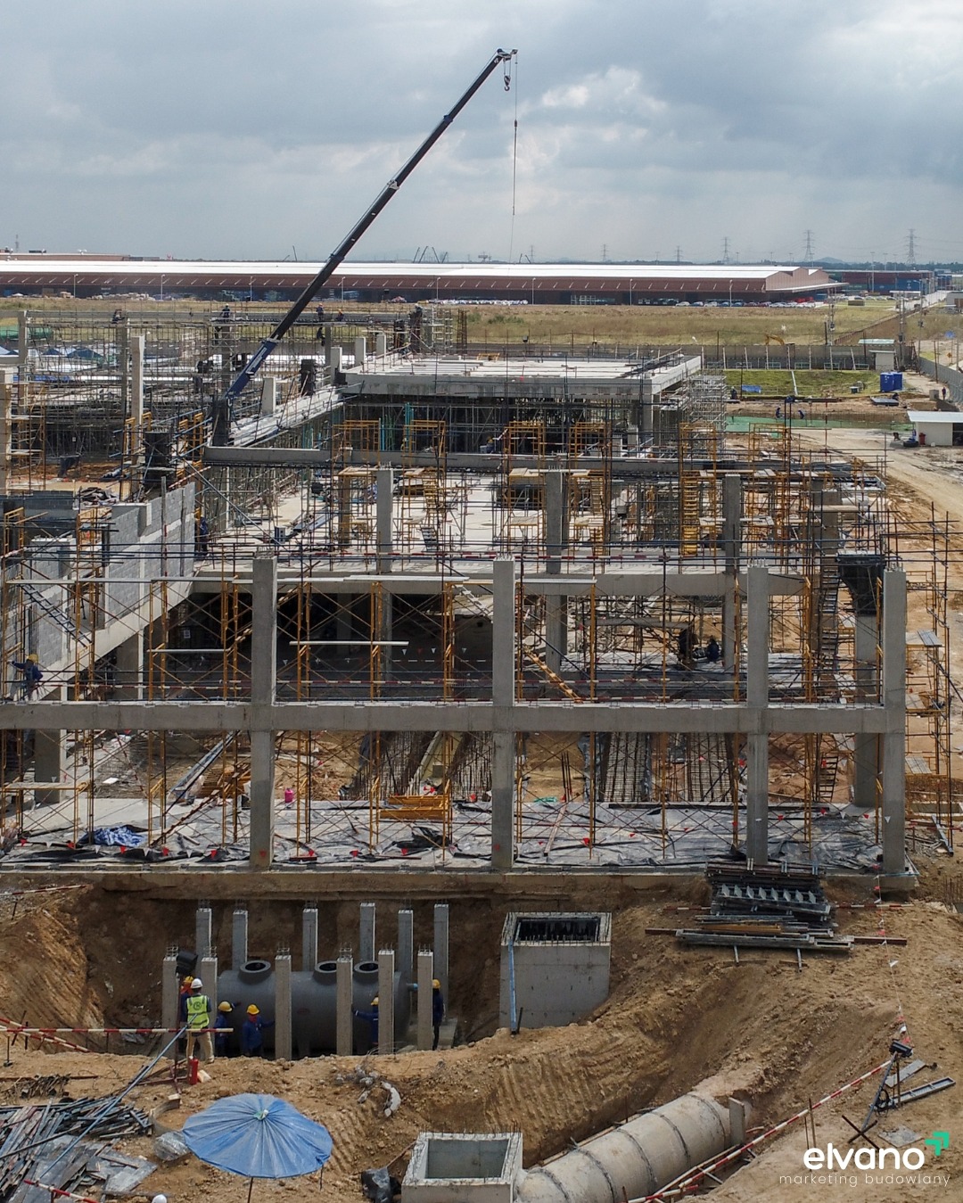 Wysoka konstrukcja budynku w stanie surowym, z licznymi rusztowaniami i dźwigiem w tle, widoczni robotnicy w kaskach przy podziemnej instalacji.